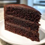 A close-up of a moist, multi-layered slice of chocolate fudge cake topped with glossy ganache.