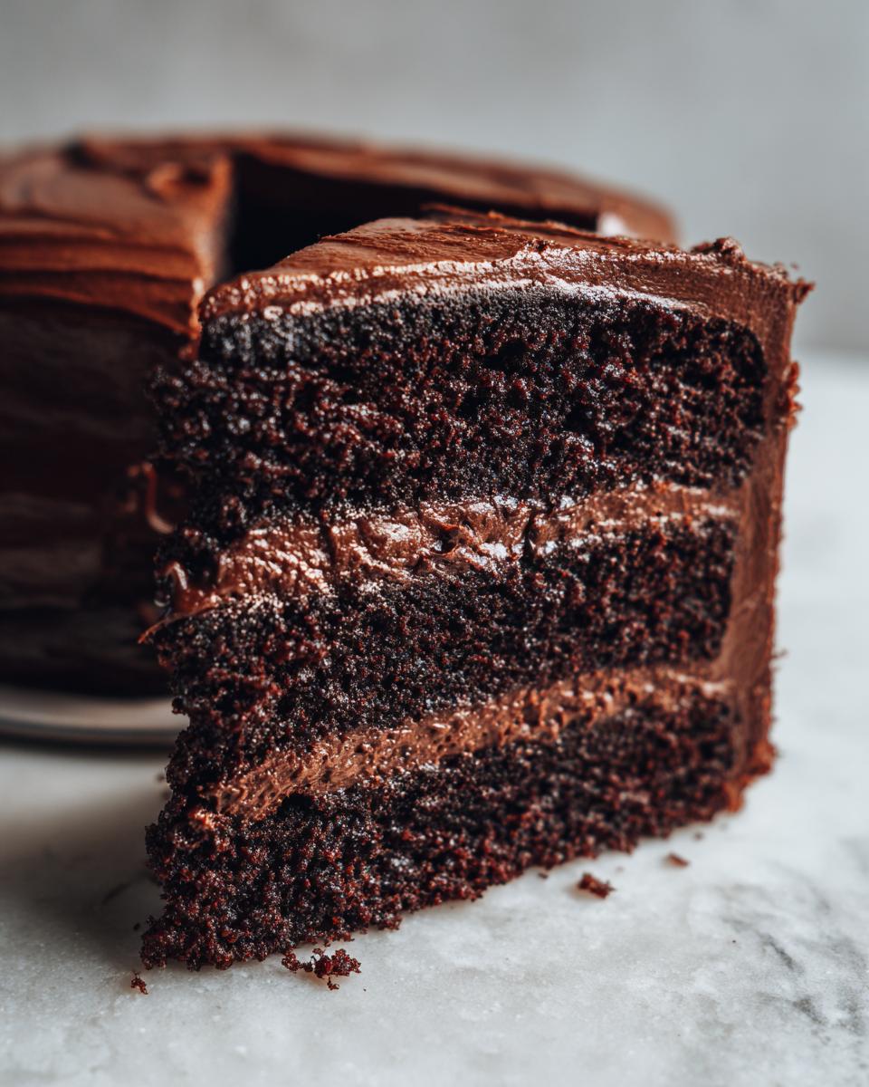 Close-up of a moist slice of three-layer chocolate fudge cake with thick chocolate frosting between layers.