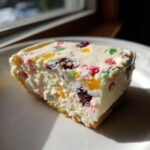 A close-up slice of white christmas tree cheesecake filled with colorful candied fruit pieces on a white plate.