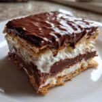 Close-up of a layered eclair cake slice featuring graham crackers, white cream, chocolate pudding, and a thick chocolate glaze.
