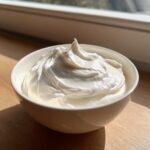 A white bowl filled with thick, perfectly peaked royal icing recipe mixture, sitting on a wooden surface near a window.
