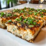 A thick, golden-brown slice of baked sushi bake casserole topped with furikake and fresh green onions.