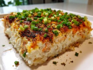 A close-up, featured image of a square slice of creamy, baked rice and seafood topping, garnished with green onions, representing the sushi bake.