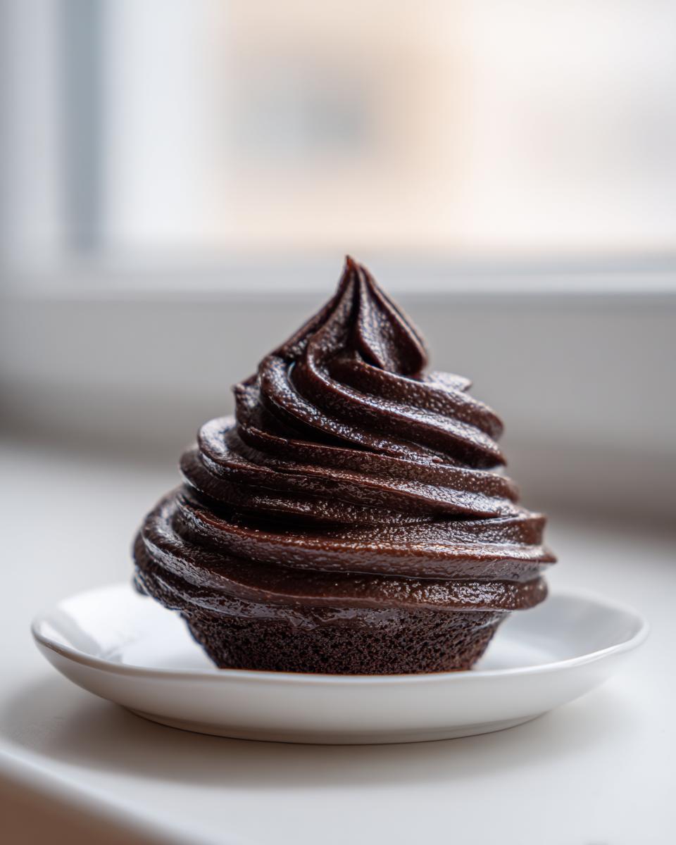 A single cupcake topped with a tall, glossy swirl of rich, dark chocolate frosting, sitting on a small white plate.