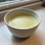 A close-up of a bowl of smooth, light green velvety celery soup resting on a light countertop near a window.