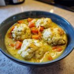 A close-up of a blue bowl filled with creamy chicken and dumplings, featuring fluffy dumplings and bright orange carrots.