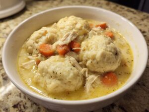 Close-up of a white bowl filled with creamy chicken and dumplings, featuring large fluffy dumplings and sliced carrots.