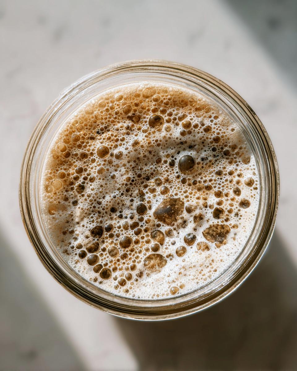 Overhead view of an active sourdough starter showing many bubbles and foam inside a glass jar.