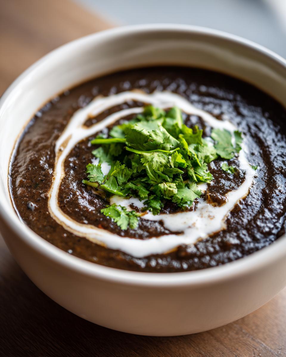 Close-up of a bowl of rich, dark black bean soup garnished with a swirl of cream and fresh cilantro.