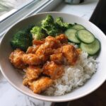 A white bowl filled with white rice topped with saucy Bang Bang Chicken, steamed broccoli, and sliced cucumbers.