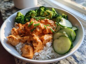 A white bowl filled with white rice topped with creamy orange Bang Bang Chicken, steamed broccoli, and sliced cucumbers.