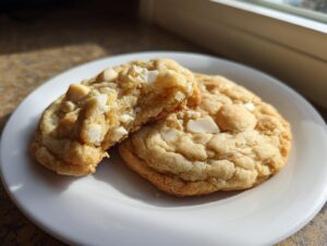 Two soft, golden macadamia nut cookies on a white plate, one broken open showing the chewy interior.