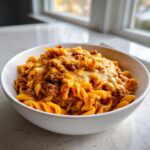 A white bowl filled with cheesy taco pasta, featuring ground beef and melted cheddar cheese topping.