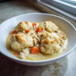A white bowl filled with creamy chicken and dumpling stew, featuring fluffy dumplings and shredded chicken.