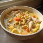 Close-up of a bowl of creamy chicken noodle soup featuring shredded chicken, egg noodles, and sliced carrots.