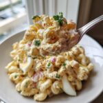 A spoonful of creamy, delicious macaroni salad featuring elbow pasta, red onion, and parsley, lifted above a bowl.