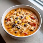 A close-up of a white bowl filled with creamy slow cooker buffalo chicken chili, topped with shredded cheese, black beans, and corn.