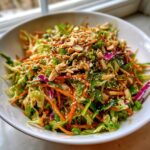 A vibrant bowl of crunchy cabbage salad mixed with shredded carrots and topped generously with chopped peanuts and sesame seeds.