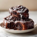 Three rich, dark chocolate cinnamon rolls stacked and drizzled with white icing on a small white plate.