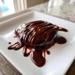 A dessert covered in thick, glossy homemade chocolate sauce dripping onto a white square plate.
