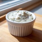 A close-up of creamy homemade tartar sauce, flecked with herbs, served in a white ribbed ramekin.
