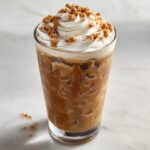 A tall glass of iced cookie butter latte topped with whipped cream and cookie crumbles.
