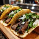 Close-up of three delicious steak tacos filled with grilled steak, diced white onion, and fresh cilantro on a wooden board.