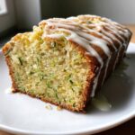 A slice of moist lemon zucchini bread topped with a thick white glaze drizzle, showing flecks of green zucchini.
