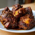 Close-up of several pieces of rich, dark brown stew chicken coated in savory sauce resting on a white plate.