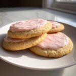 Four soft sugar cookies topped with thick, pale pink frosting stacked on a white plate.