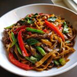 Close-up of a white bowl filled with glossy thai drunken noodles, beef strips, red peppers, and green basil leaves.