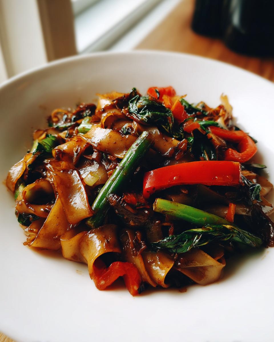 Close-up of glossy, wide rice noodles mixed with green vegetables and bright red bell peppers in thai drunken noodles.