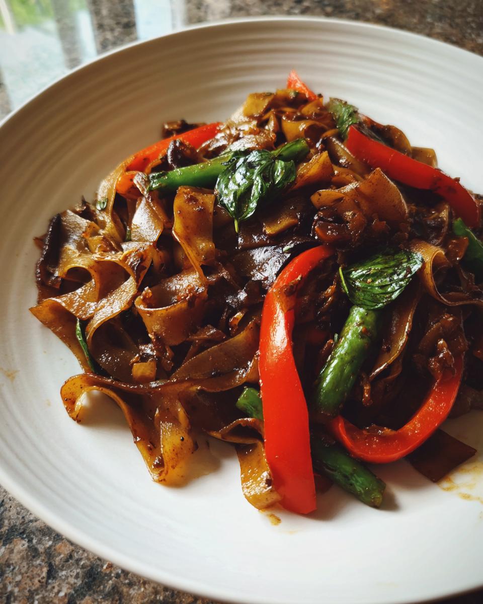 A close-up serving of savory thai drunken noodles featuring wide, dark noodles, bright red bell peppers, and green basil leaves.