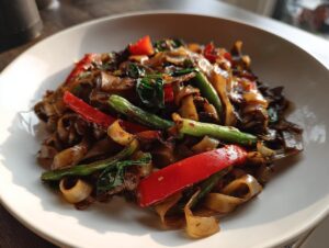 A close-up shot of a serving of saucy thai drunken noodles mixed with bright red bell peppers and green beans.