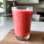 A tall glass filled with a bright pink, icy watermelon smoothie sitting on a wooden cutting board.