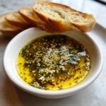 Close-up of a small white bowl filled with flavorful garlic and herb bread dip, served with toasted baguette slices.