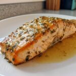 Close-up of a perfectly cooked salmon recipe fillet topped with a glistening herb and garlic glaze, sitting in pan juices.