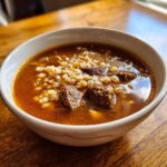 Close-up of a white bowl filled with rich, brown beef barley soup, showing chunks of beef and cooked barley grains.
