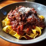A close-up of a bowl of pappardelle pasta topped generously with rich, slow-cooked beef ragu and grated Parmesan cheese.