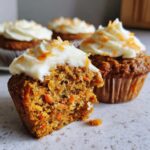 A close-up view of a bitten carrot cake muffin showing the moist interior filled with shredded carrots, topped with cream cheese frosting.