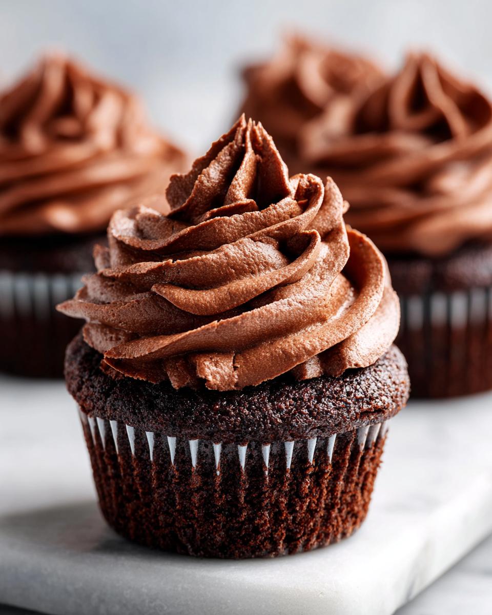A close-up of one perfectly baked chocolate cupcake topped with a swirl of rich chocolate frosting, with more chocolate cupcakes blurred in the background.