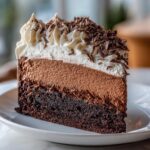 A decadent slice of chocolate mousse cake showing three distinct layers: dark cake base, light chocolate mousse, and whipped cream topping with shavings.