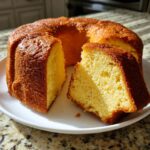 A golden brown cream cheese pound cake on a white plate with one slice cut out, showing the dense, yellow crumb.