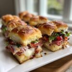 Close-up of cheesy, loaded grinder sliders on a white platter, featuring melted cheese and deli meats.