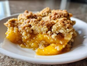 Close-up of a warm serving of peach cobbler with a thick, crumbly topping on a white plate.