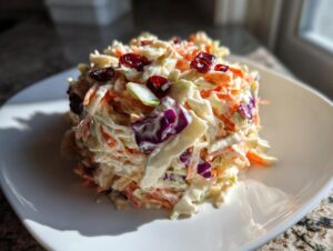 A mound of creamy apple cranberry coleslaw topped with dried cranberries, served on a white plate.