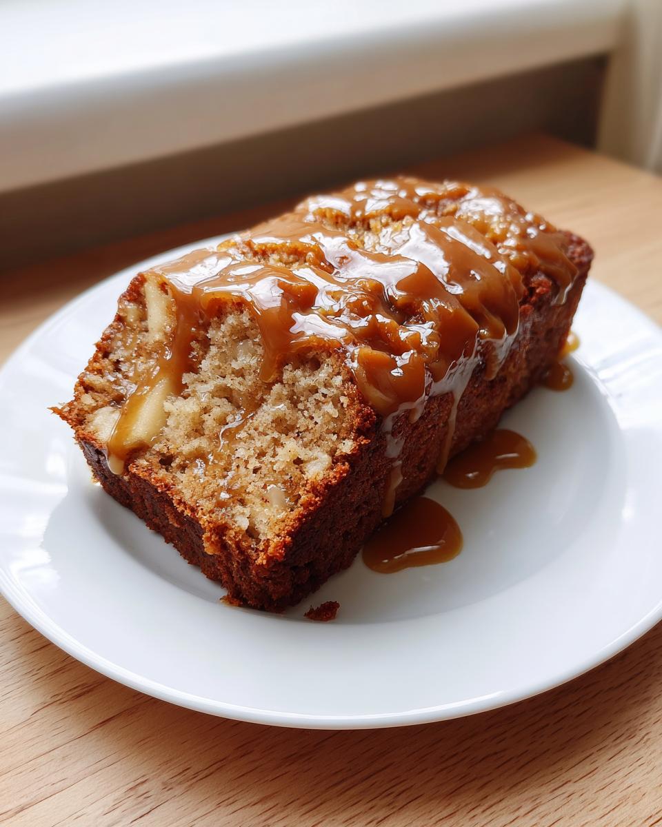 A thick slice of moist apple dapple cake topped generously with a rich caramel sauce, served on a white plate.
