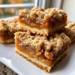 Close-up of decadent apple pie caramel apples bars with a thick caramel layer and crumb topping, stacked on a white plate.