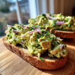 Close-up of an open-faced avocado chicken salad sandwich on whole grain bread, topped with red onion and chives.