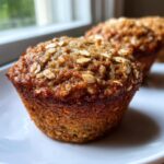 A close-up, appetizing shot of a golden brown banana oatmeal muffin topped with visible rolled oats on a white plate.